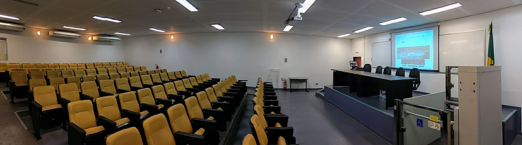 Auditório do IME, com fileiras de poltronas amarelas, mesa de honra e tela de projeção